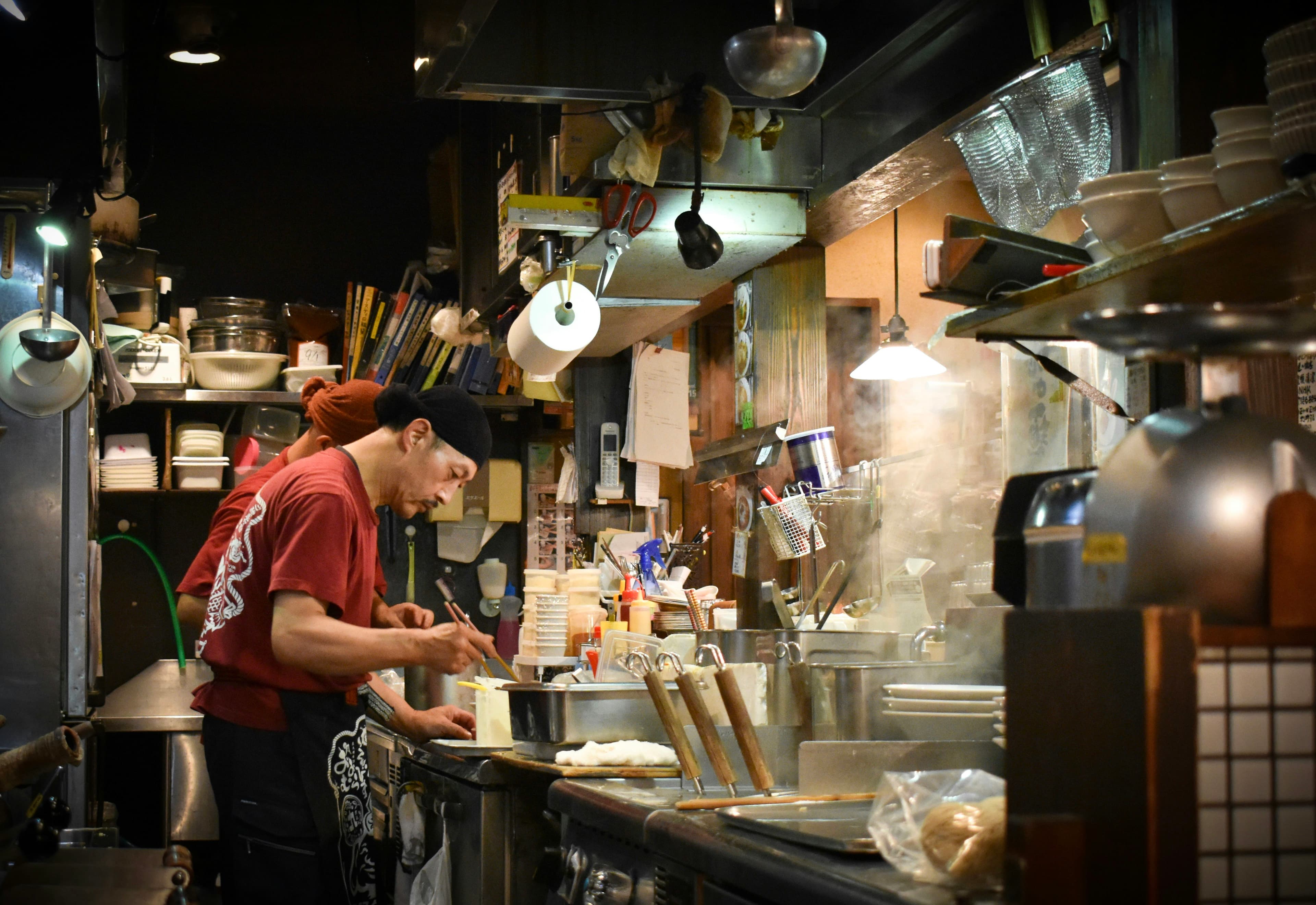 A cook working the line of a small noodle shop kitchen, bowls stacked above the pass