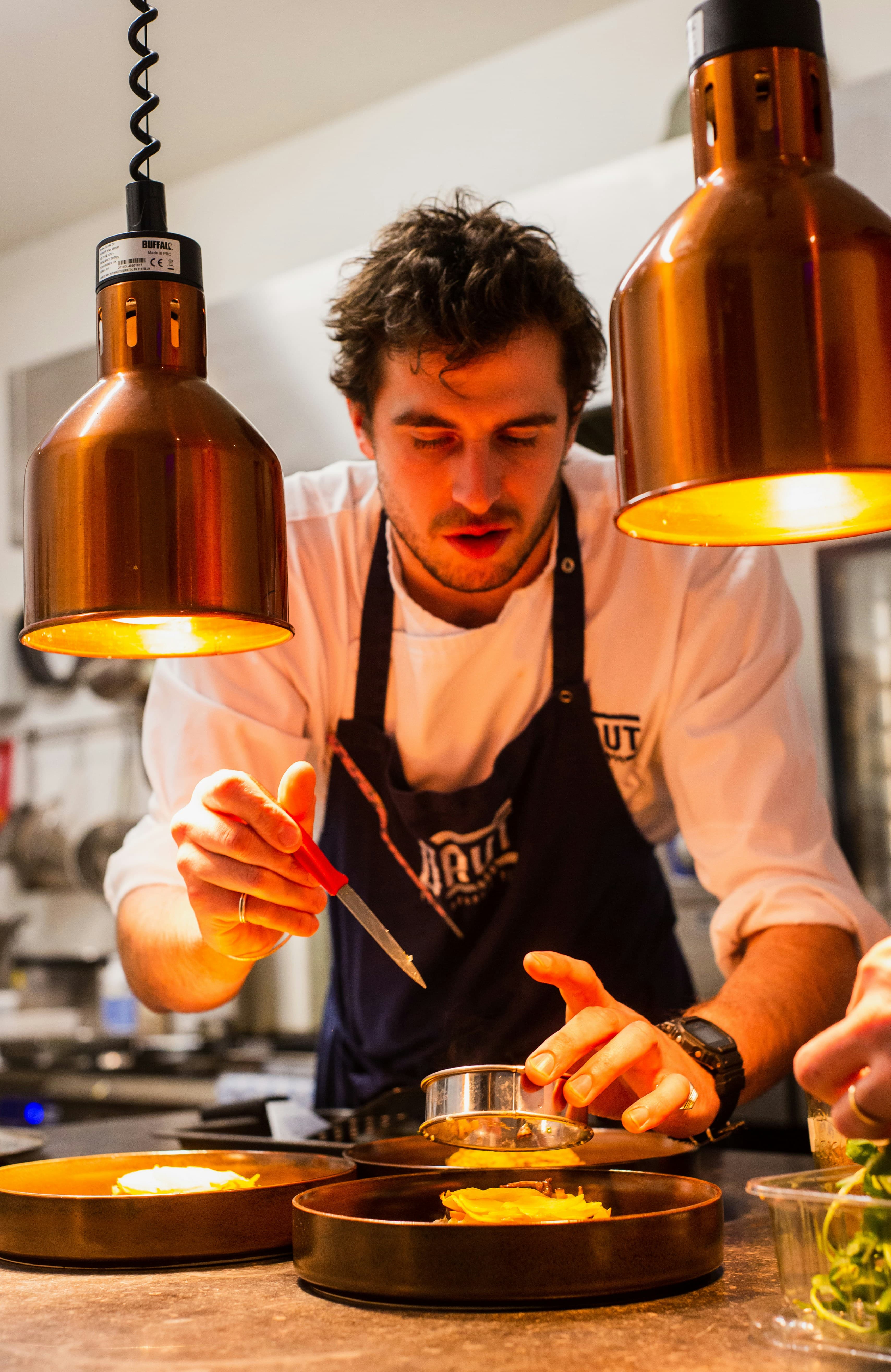 A cook in an apron plating an order under copper kitchen lamps
