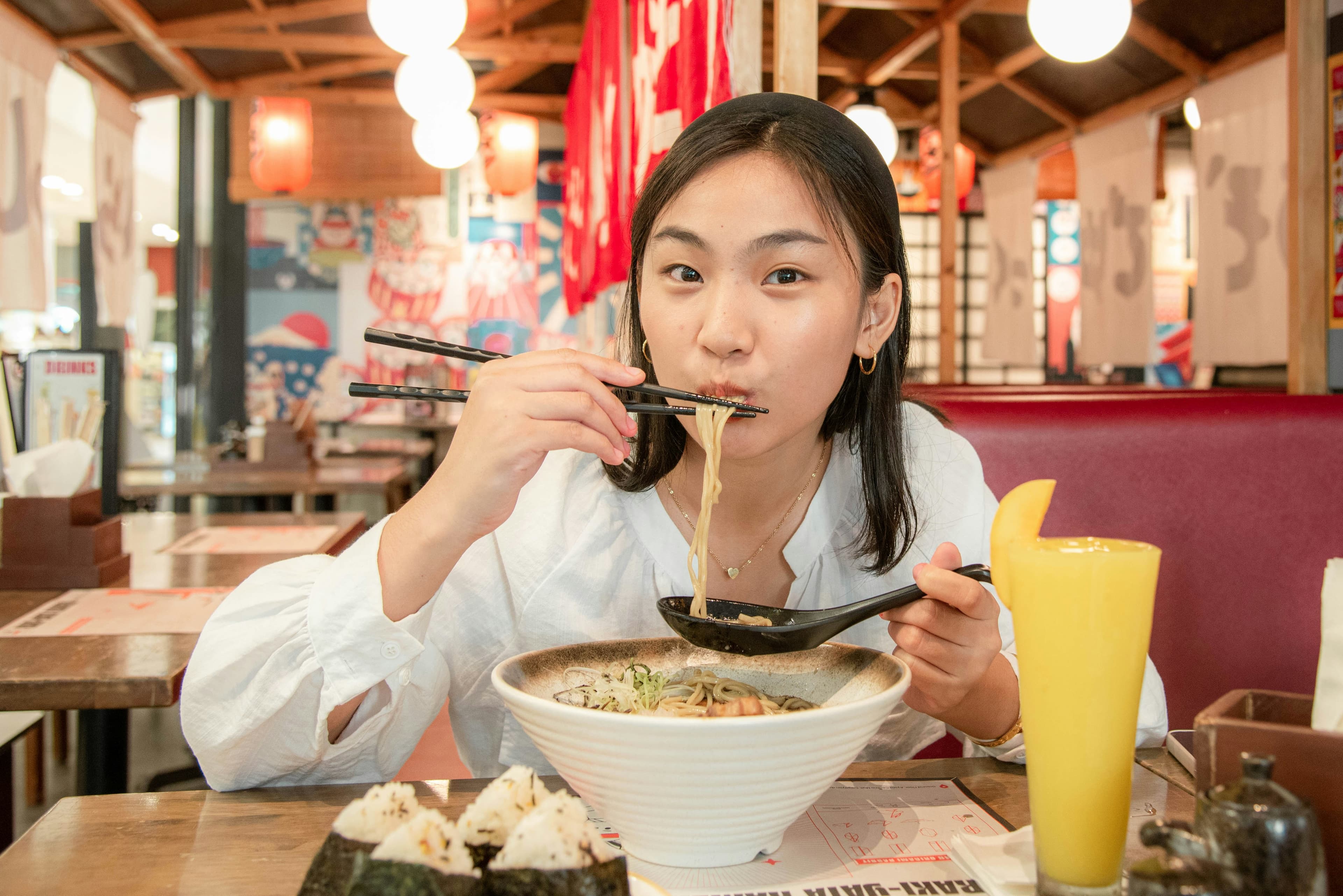A young woman eating a bowl of ramen in a busy noodle shop, chopsticks raised