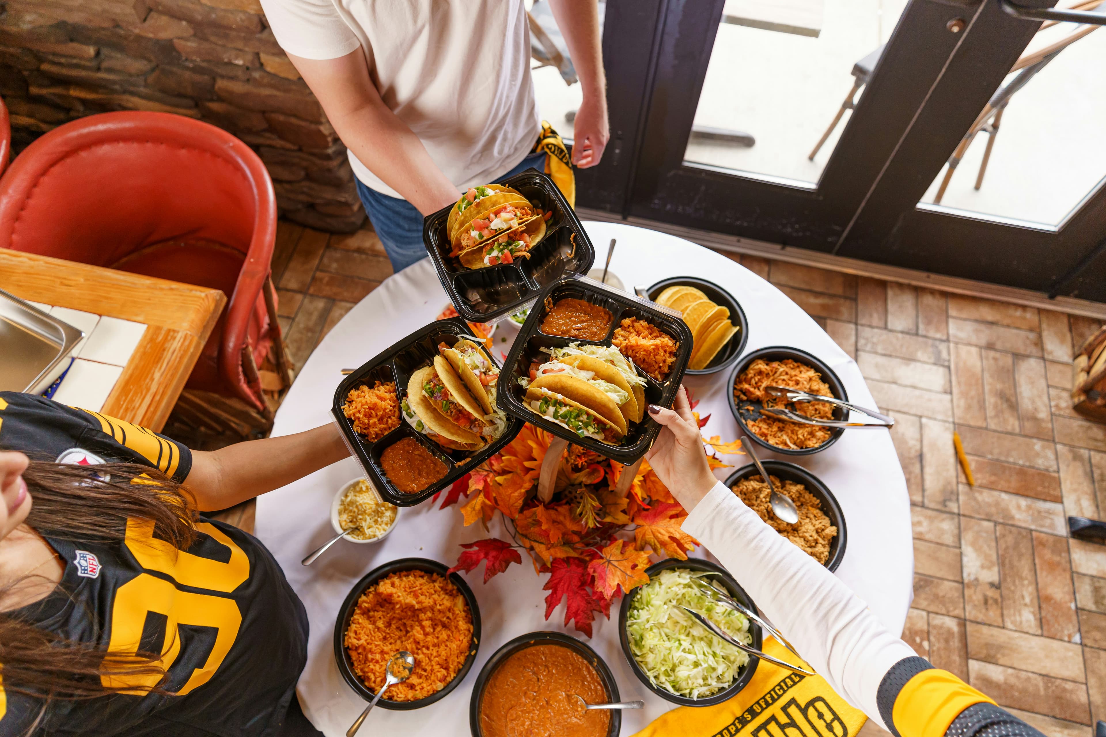 Three people reaching over a table of takeout containers of tacos, rice and beans